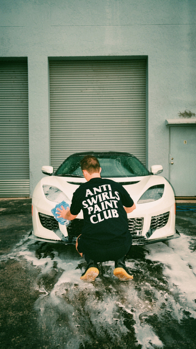 Person washing and detailing a white sports car with the Detail Driven Anti Swirls Paint Club shirt for car enthusiasts and detailers in front of a garage.